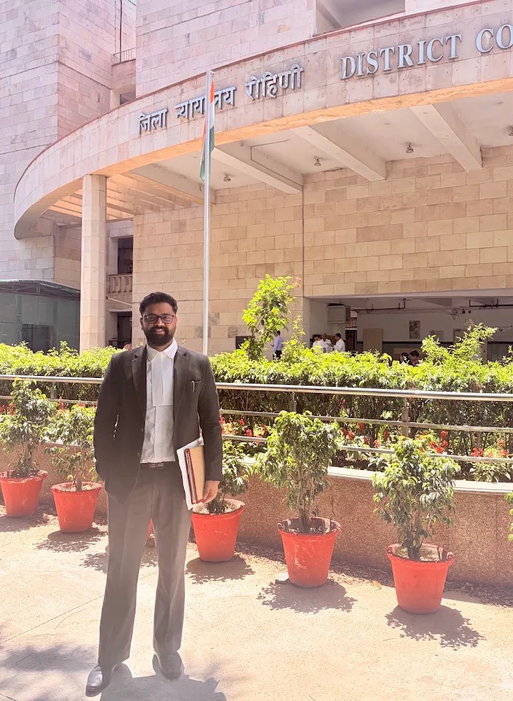 Adv. Ashutosh Hans standing outside a district court building, wearing advocate's robes and holding case files.